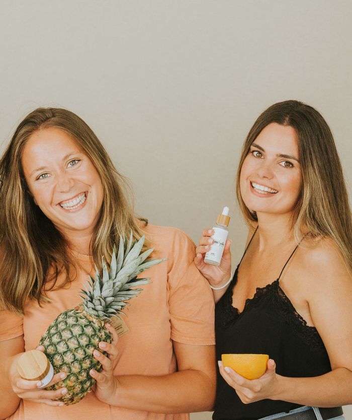 Two women holding a pineapple and skincare products, smiling against a neutral background.