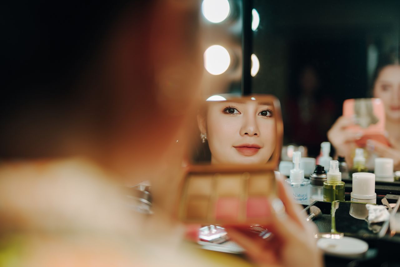 A woman applying makeup, captured in a mirror reflection surrounded by beauty products.