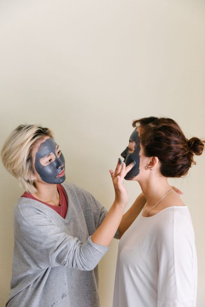 Young asian female in casual wear with cosmetic mask on face applying black facial mud mask to face of young woman on beige background